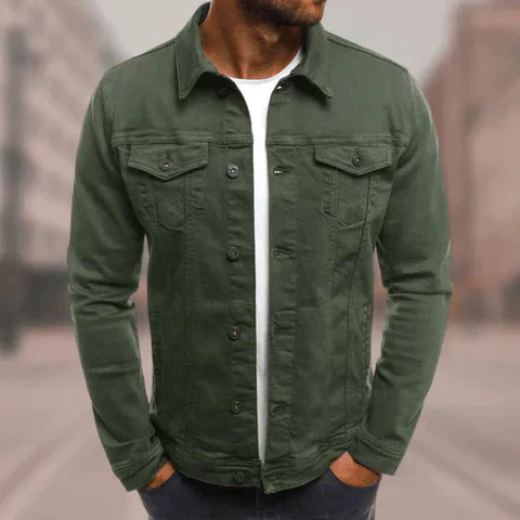 Mann in olivgrüner Jeansjacke, weißem T-Shirt, Hände in den Taschen, urbaner Hintergrund. Modische Herrenbekleidung, lässiger Stil.