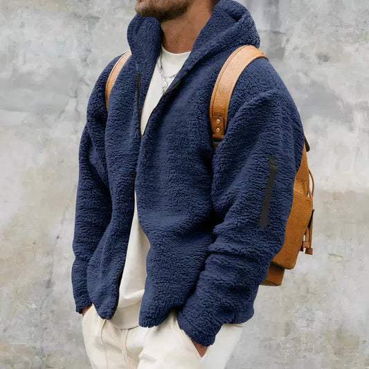Mann in blauer Fleecejacke mit Kapuze und braunem Rucksack vor grauem Hintergrund. Wintermode, Herrenbekleidung, Outdoor-Look.