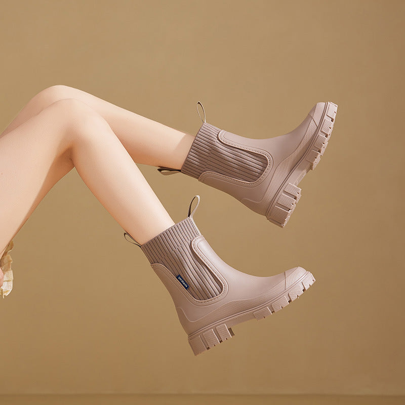 Beige Gummistiefel mit dicker Sohle und geripptem Schaft, getragen von einer Person mit überkreuzten Beinen vor beigem Hintergrund.