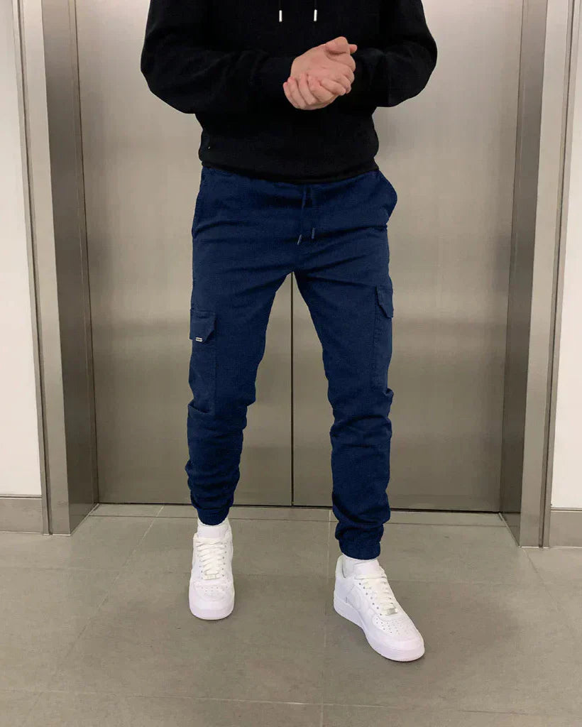 Person in blauer Jogginghose und weißen Sneakers vor einem Aufzug. Freizeitmode, Herrenbekleidung, sportlicher Look.