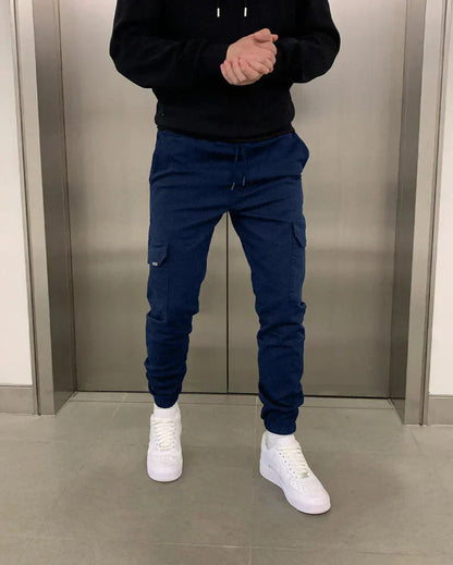 Person in blauer Jogginghose und weißen Sneakers vor einem Aufzug. Freizeitmode, Herrenbekleidung, sportlicher Look.