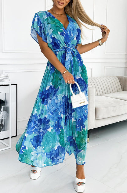 Frau in blauem, geblümtem Maxikleid mit Gürtel, hält weiße Handtasche. Elegantes Sommeroutfit, modisch, stilvoll, ideal für besondere Anlässe.