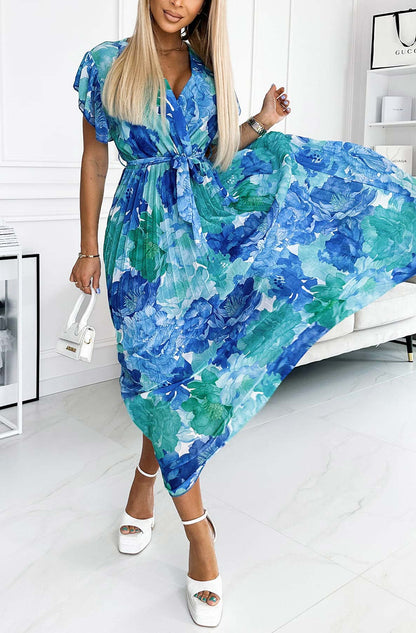 Frau in blau-grünem Blumenkleid, hält weißen Mini-Handtasche, trägt weiße Plateau-Sandalen. Elegante Sommermode, florales Design, stilvoller Look.