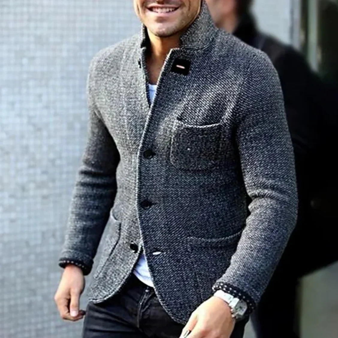 Ein Mann trägt eine graue Tweed-Jacke mit Knöpfen, kombiniert mit einem weißen Hemd und einer schwarzen Hose. Elegante Herrenmode, stilvoller Look.