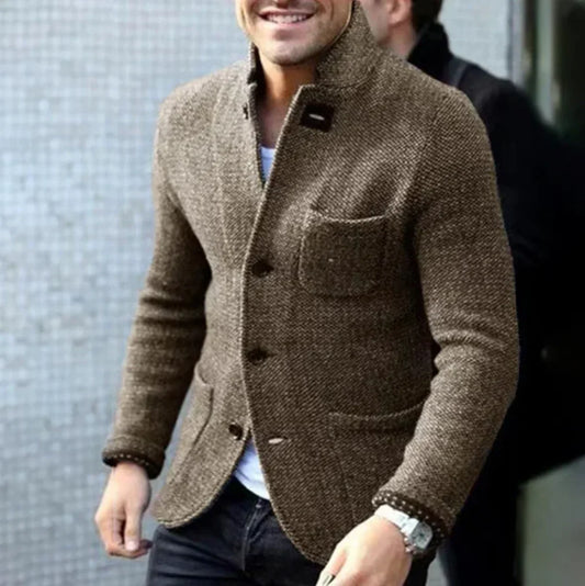 Ein Mann trägt eine braune Tweedjacke mit Knöpfen und Brusttasche, kombiniert mit einem weißen Hemd und einer schwarzen Hose. Elegante Herrenmode.