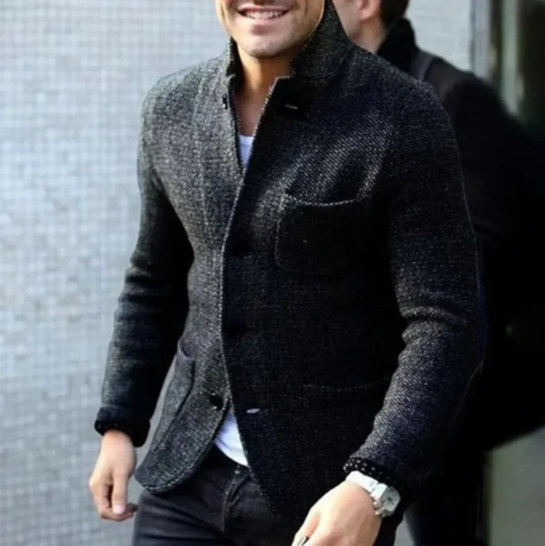 Ein Mann in einem eleganten, grauen Wollblazer mit weißem Hemd und Armbanduhr, lächelnd, vor einer modernen, strukturierten Wand.