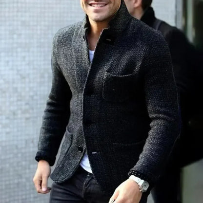 Ein Mann in einem eleganten, grauen Wollblazer mit weißem Hemd und Armbanduhr, lächelnd, vor einer modernen, strukturierten Wand.