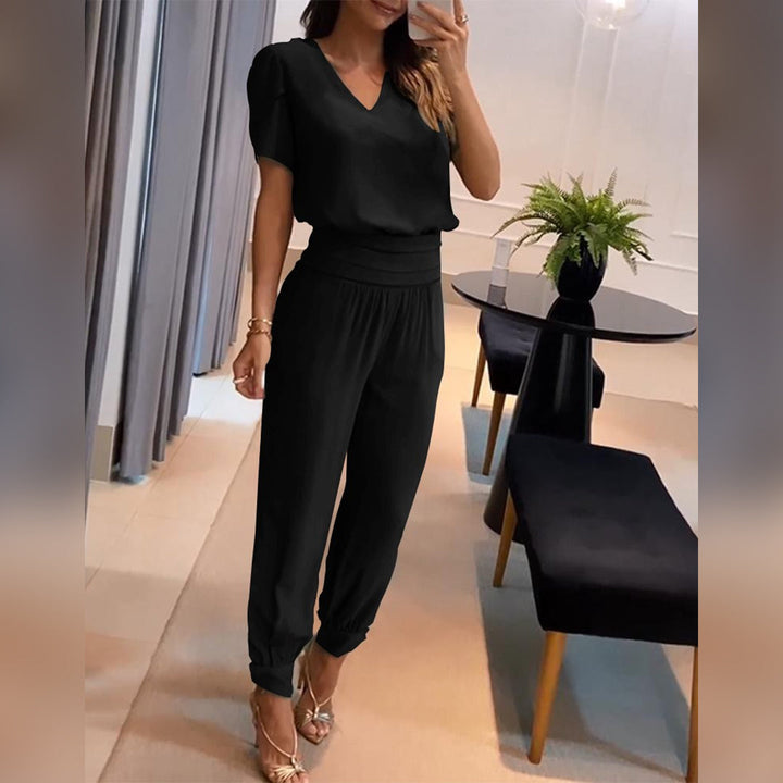 Frau in elegantem schwarzen Jumpsuit posiert in modernem Wohnzimmer, trägt goldene Sandalen, neben Tisch mit Pflanze. Mode, Damenbekleidung, Stil.
