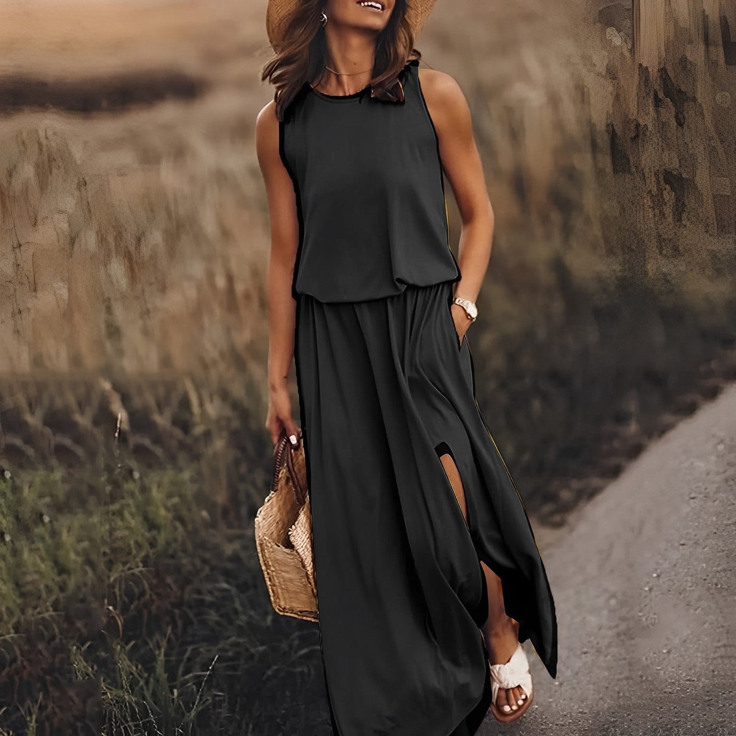 Frau in schwarzem Maxikleid und Strohhut spaziert auf ländlichem Weg. Sommermode, elegantes Outfit, Freizeitkleidung, Naturkulisse.