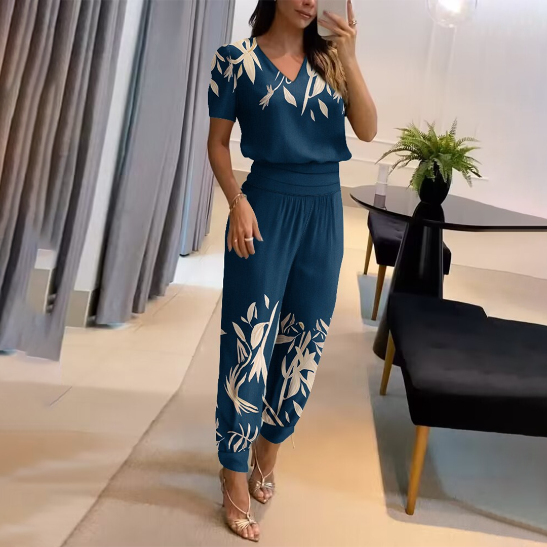 Frau in blauem, geblümtem Jumpsuit posiert in modernem Wohnzimmer. Elegante Mode, stilvolle Inneneinrichtung, trendige Damenbekleidung.