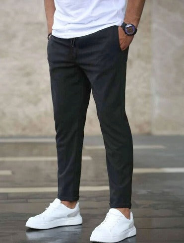 Mann in schwarzer Jogginghose und weißen Sneakers, trägt weißes T-Shirt, steht auf grauem Boden. Lässige Herrenmode, Streetwear, Freizeitlook.