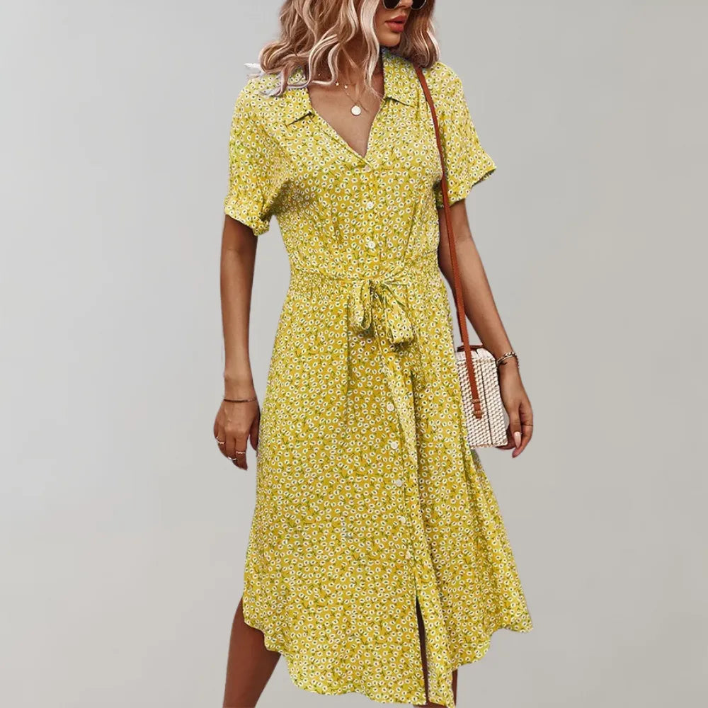 Frau in gelbem Sommerkleid mit Blumenmuster, Bindegürtel und V-Ausschnitt, trägt eine Umhängetasche. Modetrend Damenkleider 2023.