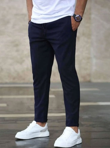 Mann in dunkelblauer Chinohose und weißen Sneakern, trägt weißes T-Shirt, steht auf grauem Boden. Herrenmode, lässiger Stil, moderne Kleidung.