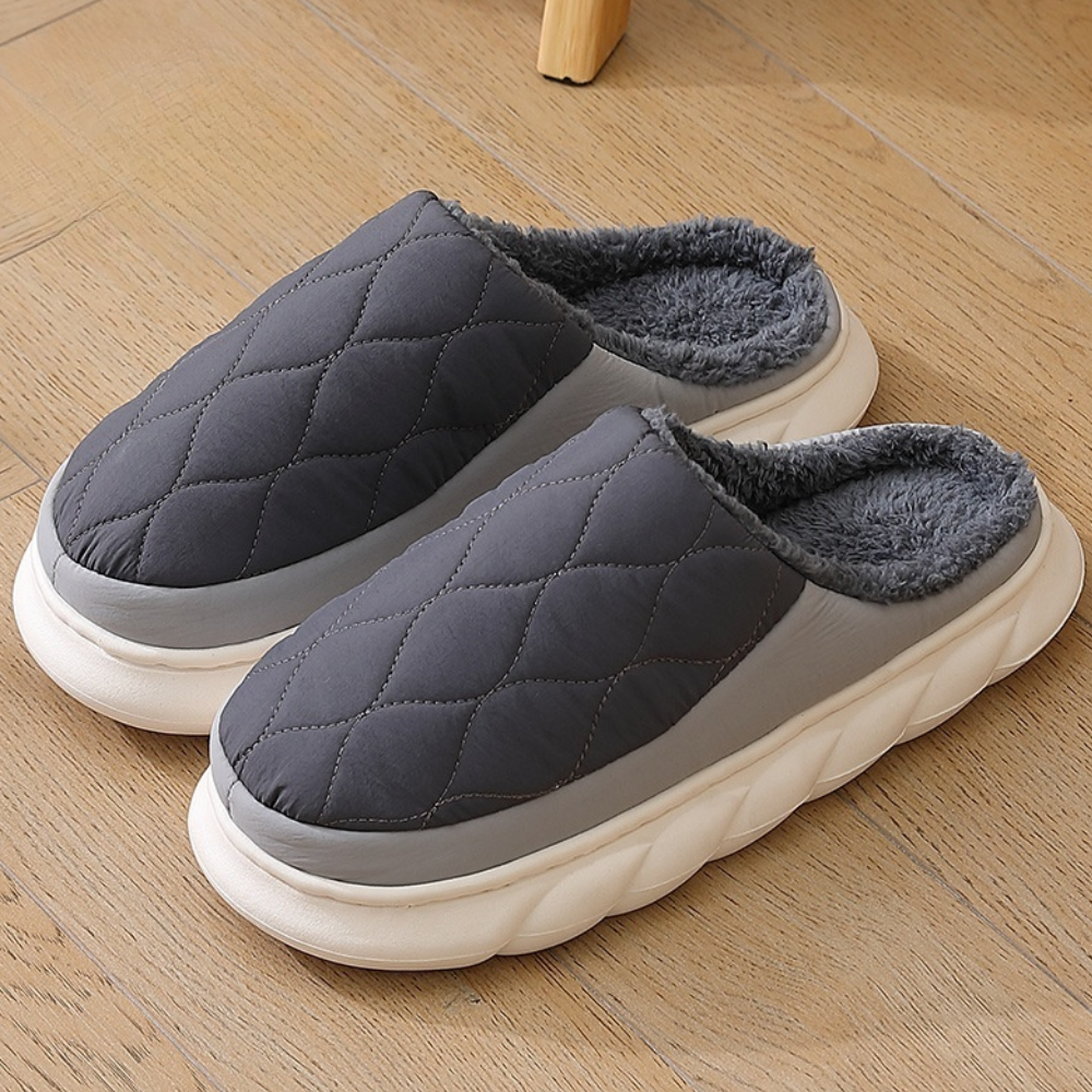 Graue, gesteppte Hausschuhe mit flauschigem Innenfutter auf Holzboden. Bequeme, warme Pantoffeln für Zuhause. Ideal für kalte Wintertage.