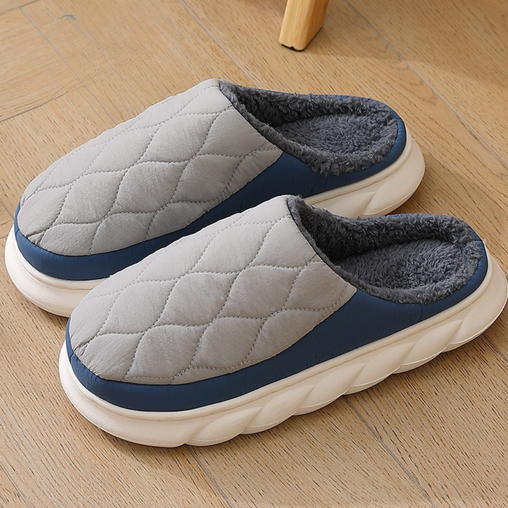 Graue, gesteppte Hausschuhe mit blauer Umrandung und flauschigem Innenfutter auf Holzboden. Bequeme, warme Pantoffeln für Zuhause.