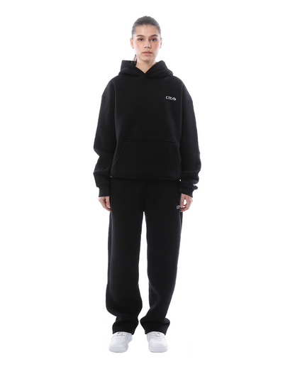 Frau in schwarzem Hoodie und Jogginghose, steht frontal, weiße Turnschuhe, minimalistischer Stil, Freizeitmode, isolierter weißer Hintergrund.