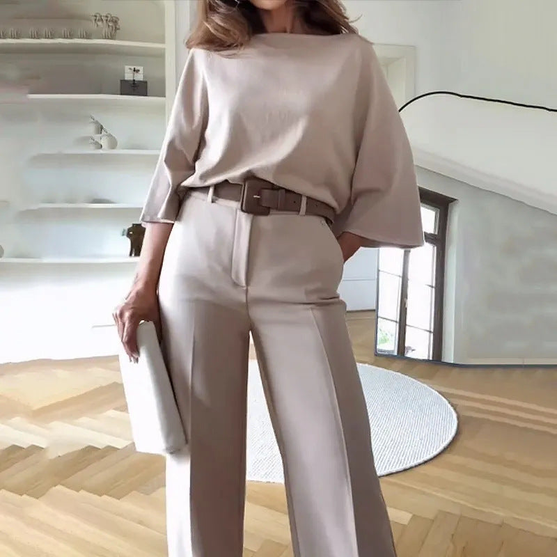 Frau in eleganter, beiger Kleidung mit weitem Oberteil und Hose, hält eine Clutch. Modernes Wohnzimmer im Hintergrund. Minimalistischer Stil.