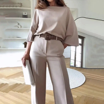 Frau in eleganter, beiger Kleidung mit weitem Oberteil und Hose, hält eine Clutch. Modernes Wohnzimmer im Hintergrund. Minimalistischer Stil.