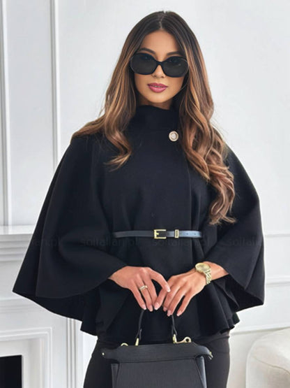 Frau in schwarzem Cape mit Gürtel, Sonnenbrille und Handtasche, steht vor weißer Wand. Elegante Mode, stilvolles Outfit, modernes Accessoire.