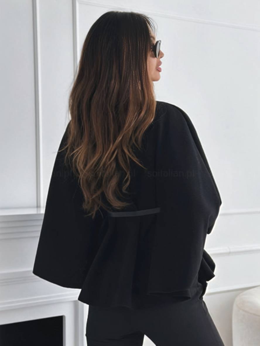 Frau mit langen braunen Haaren, trägt schwarze Sonnenbrille und eleganten schwarzen Blazer, steht vor weißer Wand. Fashion, Stil, Mode.