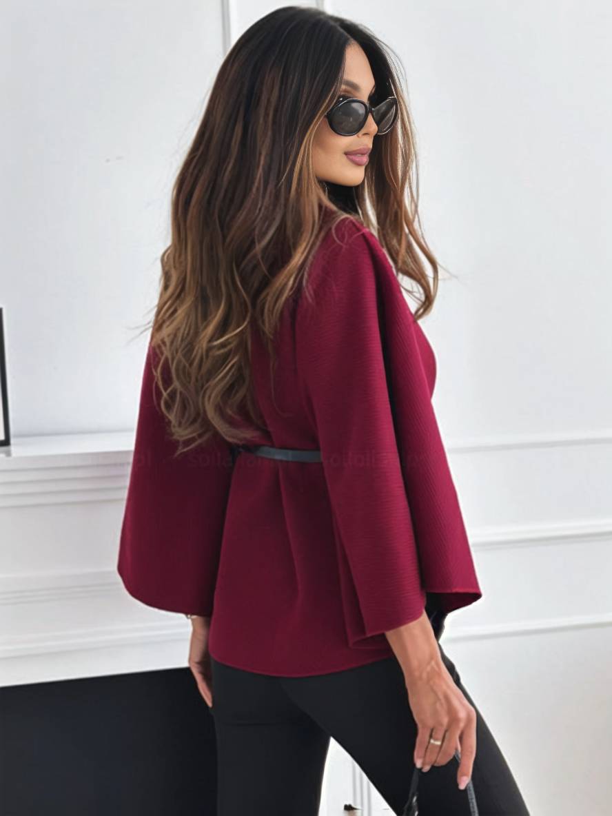 Frau mit langen Haaren in rotem Blazer und schwarzer Hose, trägt Sonnenbrille, steht vor weißer Wand. Elegante Mode, stilvolles Outfit.