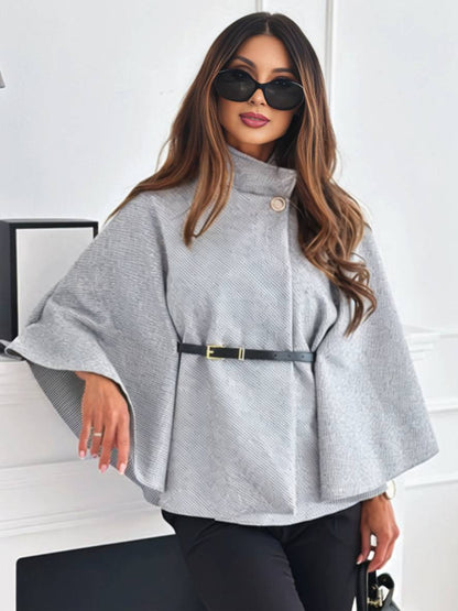 Frau in grauem Poncho mit Gürtel, Sonnenbrille und langen Haaren, steht vor weißer Wand. Elegante Herbstmode, stilvoller Look.