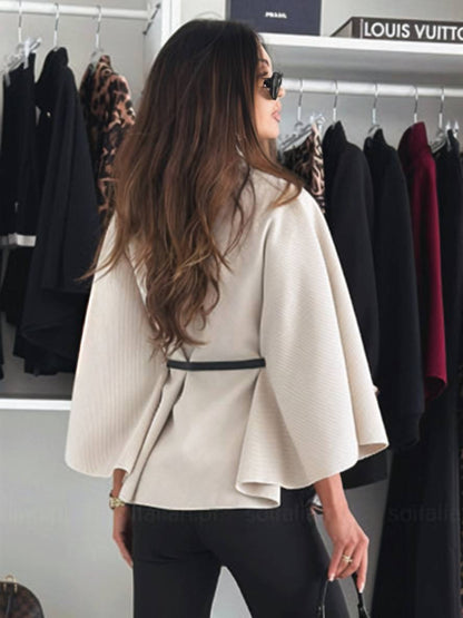 Frau in stilvollem, beigem Cape mit Gürtel, Sonnenbrille, vor Kleiderschrank mit eleganter Kleidung. Mode, Damenbekleidung, Herbsttrends.