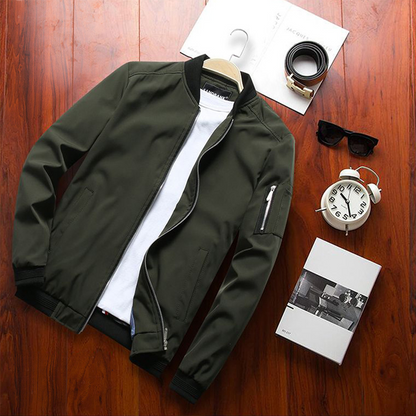 Olivgrüne Bomberjacke auf Holzboden, kombiniert mit weißem T-Shirt, schwarzer Sonnenbrille, Gürtel, Wecker und Buch. Herrenmode, stilvoll, modern.