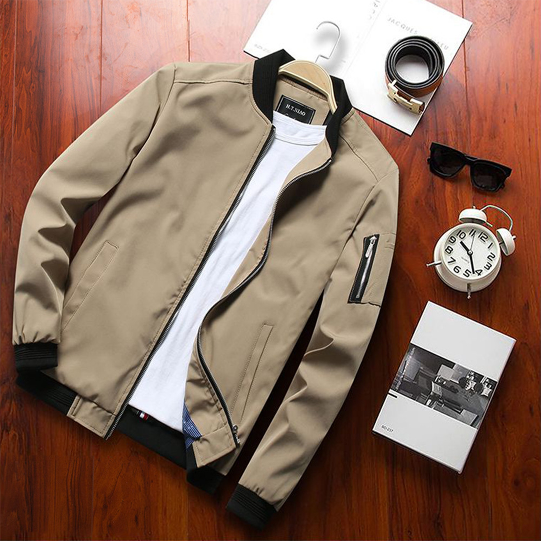Beige Herren-Bomberjacke auf Holztisch, kombiniert mit weißem T-Shirt, Uhr, Sonnenbrille und Parfüm. Modische Herrenbekleidung Accessoires.