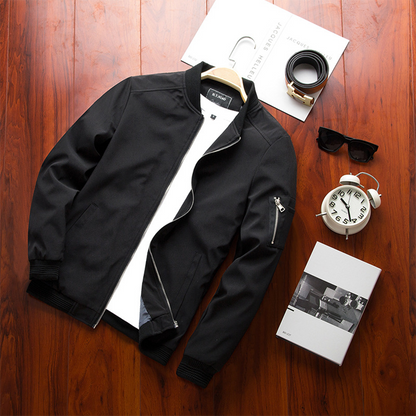 Schwarze Bomberjacke mit weißem T-Shirt auf Holzboden, umgeben von Armbanduhr, Sonnenbrille, Wecker und Buch. Herrenmode, stilvolle Accessoires.