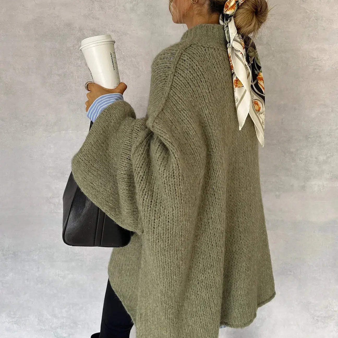 Frau in oversized Strickpullover, hält Kaffeebecher und schwarze Tasche, trägt Haarband mit Muster. Herbstmode, lässiger Stil, Streetwear.