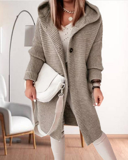 Frau in grauem Strickmantel mit Kapuze, weißer Handtasche und weißem Outfit in modernem Wohnzimmer. Modetrend, Herbstmode, Damenbekleidung.