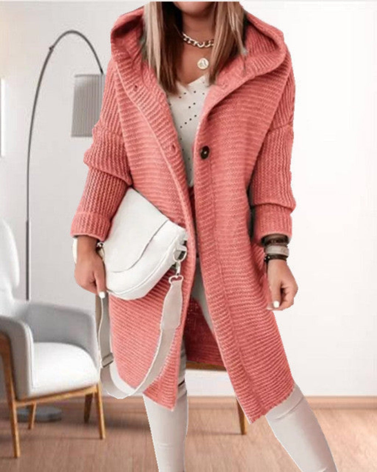 Frau in rosa Strickmantel mit Kapuze, weißer Tasche und heller Kleidung. Moderne Mode, gemütlicher Look, Innenraum mit Stuhl und Lampe im Hintergrund.