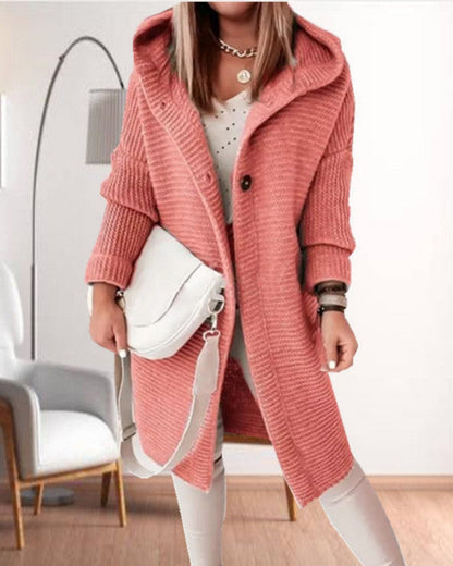 Frau in rosa Strickmantel mit Kapuze, weißer Tasche und heller Kleidung. Moderne Mode, gemütlicher Look, Innenraum mit Stuhl und Lampe im Hintergrund.