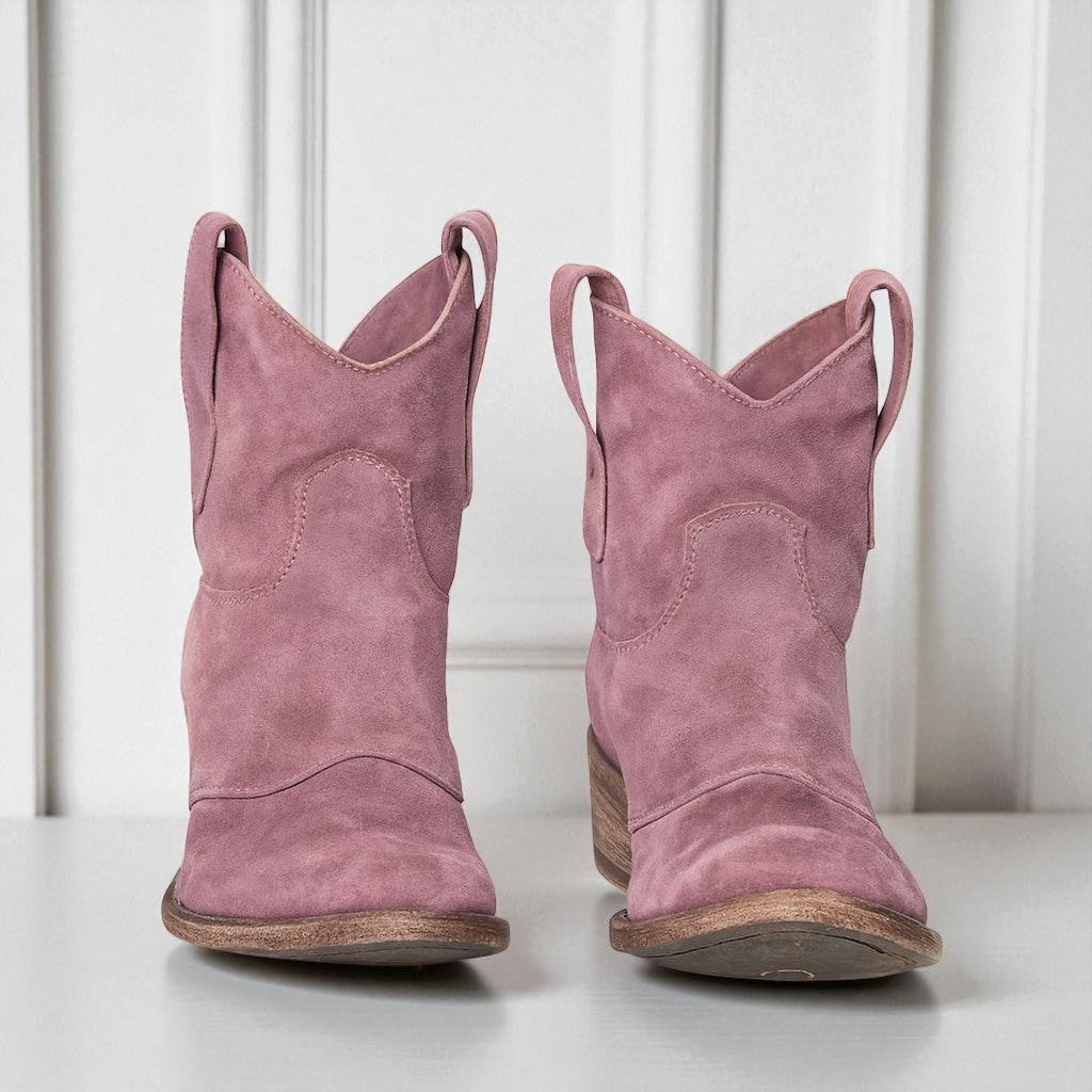 Rosa Wildleder-Cowboystiefel mit Holzabsatz vor weißer Wand. Modische Damenstiefel, Westernstil, ideal für Herbstmode und lässige Outfits.