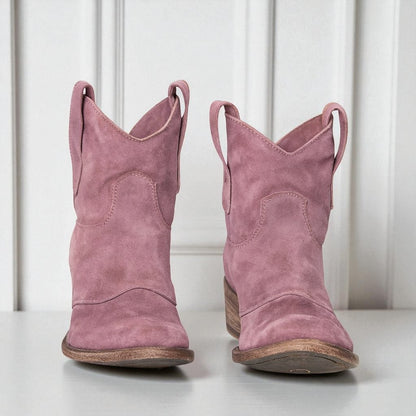 Rosa Wildleder-Cowboystiefel mit Holzabsatz vor weißer Wand. Modische Damenstiefel, Westernstil, ideal für Herbstmode und lässige Outfits.