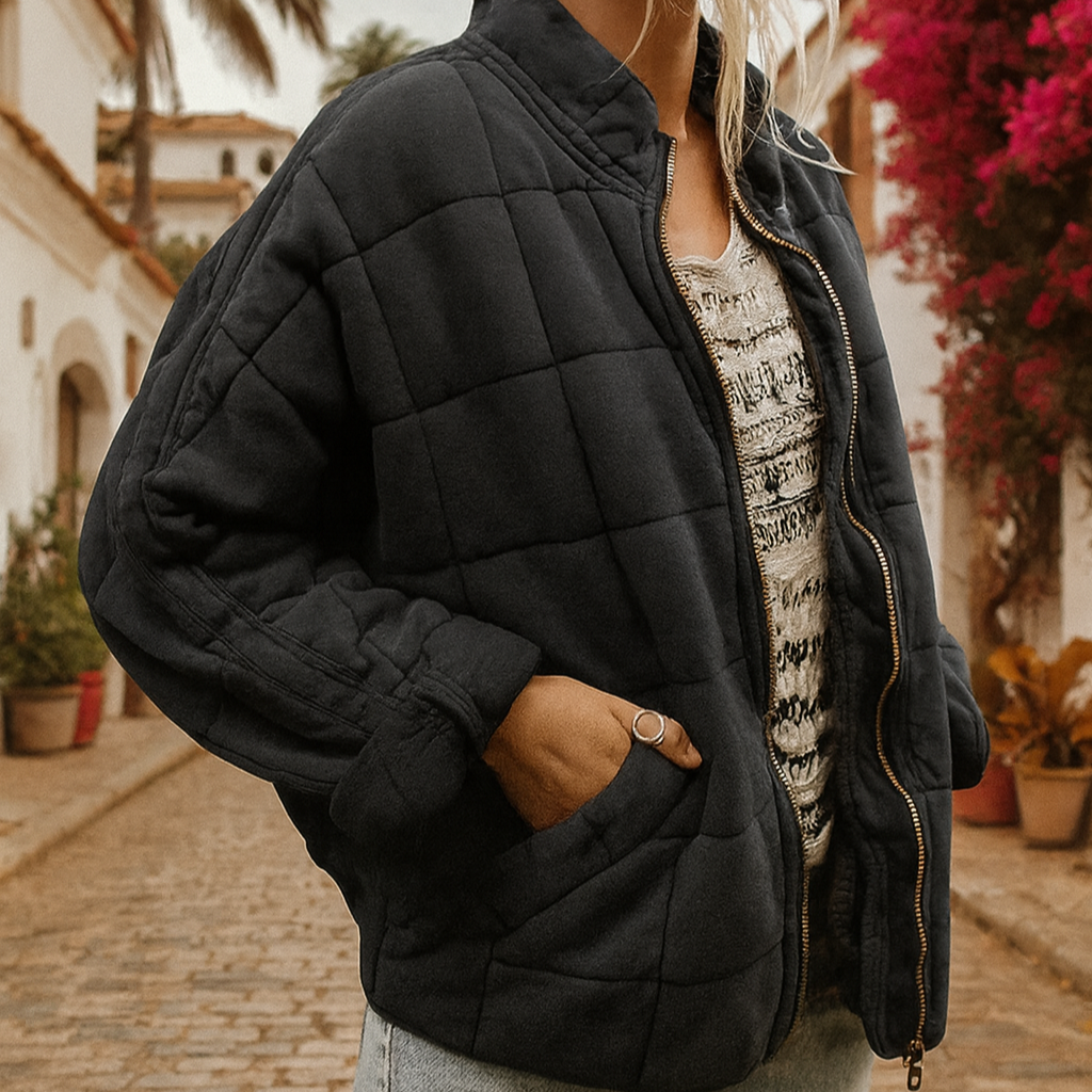 Frau in schwarzer Steppjacke auf gepflasterter Straße, umgeben von blühenden Bougainvillea und mediterraner Architektur. Mode, Herbst, Outdoor.
