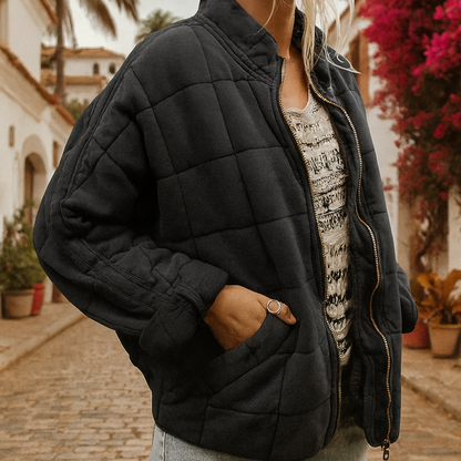 Frau in schwarzer Steppjacke auf gepflasterter Straße, umgeben von blühenden Bougainvillea und mediterraner Architektur. Mode, Herbst, Outdoor.