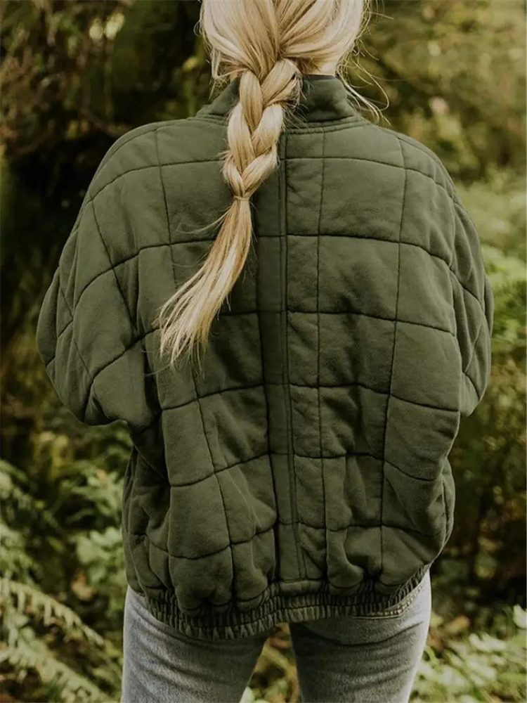 Blonde Frau mit geflochtenem Zopf trägt eine olivgrüne, gesteppte Jacke im Wald. Outdoor-Mode, Herbstbekleidung, Natur, Wanderoutfit.