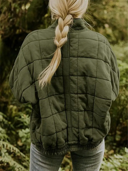 Blonde Frau mit geflochtenem Zopf trägt eine olivgrüne, gesteppte Jacke im Wald. Outdoor-Mode, Herbstbekleidung, Natur, Wanderoutfit.
