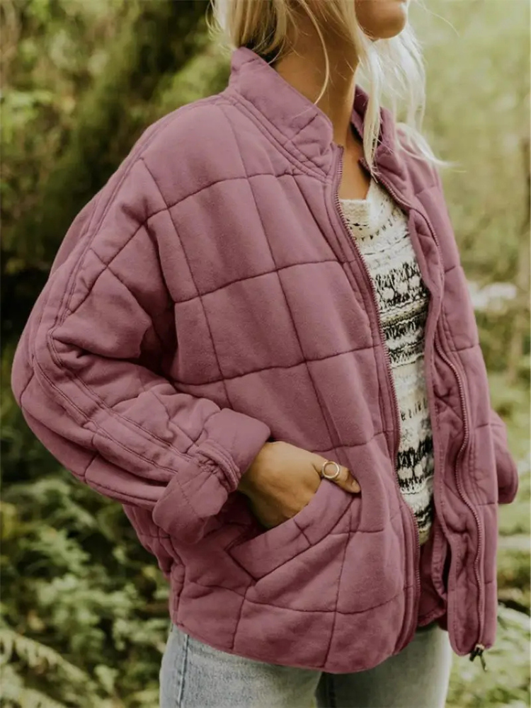 Frau in lila Steppjacke, lässig über gestreiftem Pullover, steht im Freien. Modetrend Herbst, bequeme Damenbekleidung, Outdoor-Stil.