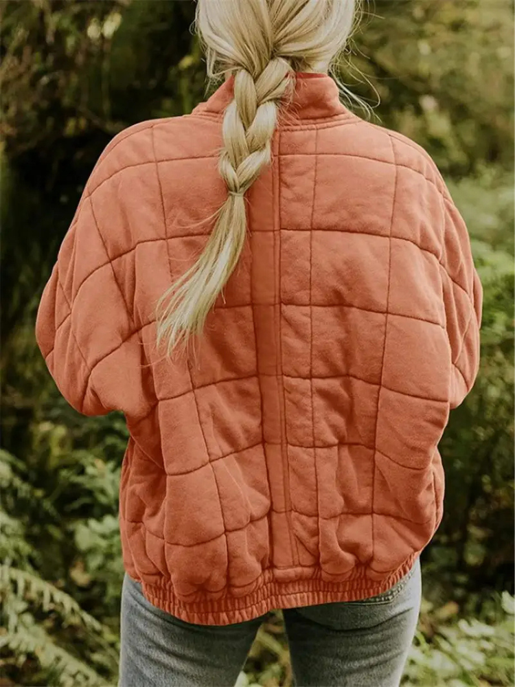 Frau mit geflochtenem Haar trägt eine orange gesteppte Jacke im Wald. Outdoor-Mode, Herbstkleidung, Natur, Freizeitkleidung.