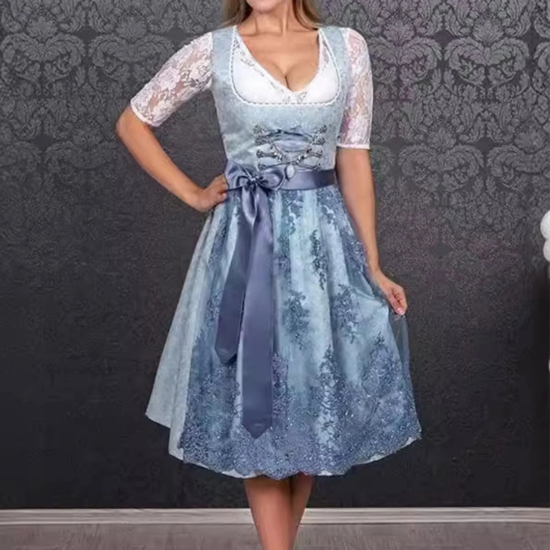 Frau in traditionellem blauen Dirndl mit Spitzenärmeln vor gemusterter Tapete. Oktoberfest Mode, bayerische Tracht, elegante Damenbekleidung.