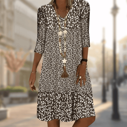 Frau in geblümtem Sommerkleid mit langen Ketten, spaziert auf sonniger Stadtstraße. Modetrend, Damenmode, Freizeitkleid, urbaner Stil.