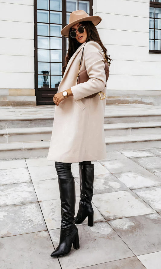 Frau in beigem Mantel und Hut, schwarze Stiefel, Sonnenbrille, braune Tasche, steht vor einem Gebäude. Elegante Herbstmode, stilvoller Look.