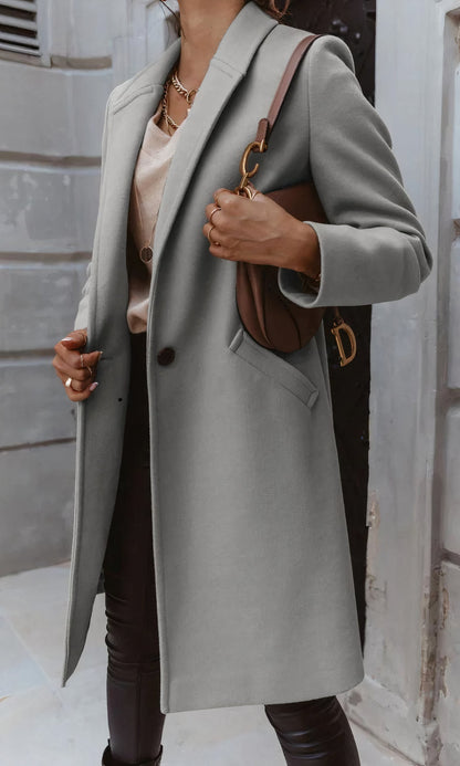 Frau in grauem Mantel, brauner Lederhandtasche, beiger Bluse und schwarzer Hose vor urbanem Hintergrund. Elegante Herbstmode, Streetstyle.