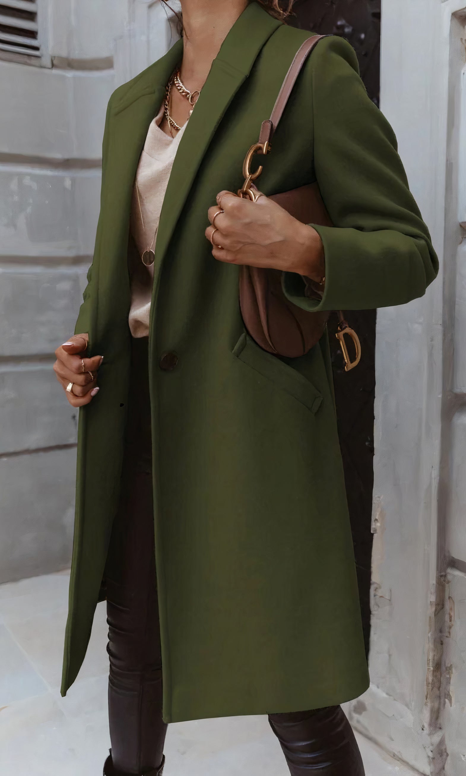 Frau in olivgrünem Mantel, hält braune Designer-Handtasche, trägt beige Bluse und schwarze Hose. Elegante Herbstmode, urbaner Hintergrund.