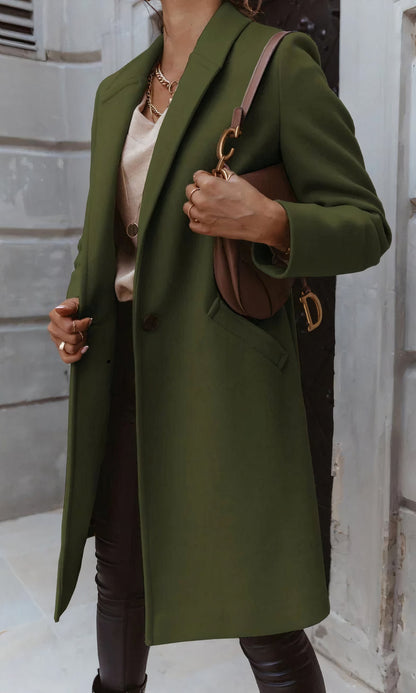 Frau in olivgrünem Mantel, hält braune Designer-Handtasche, trägt beige Bluse und schwarze Hose. Elegante Herbstmode, urbaner Hintergrund.
