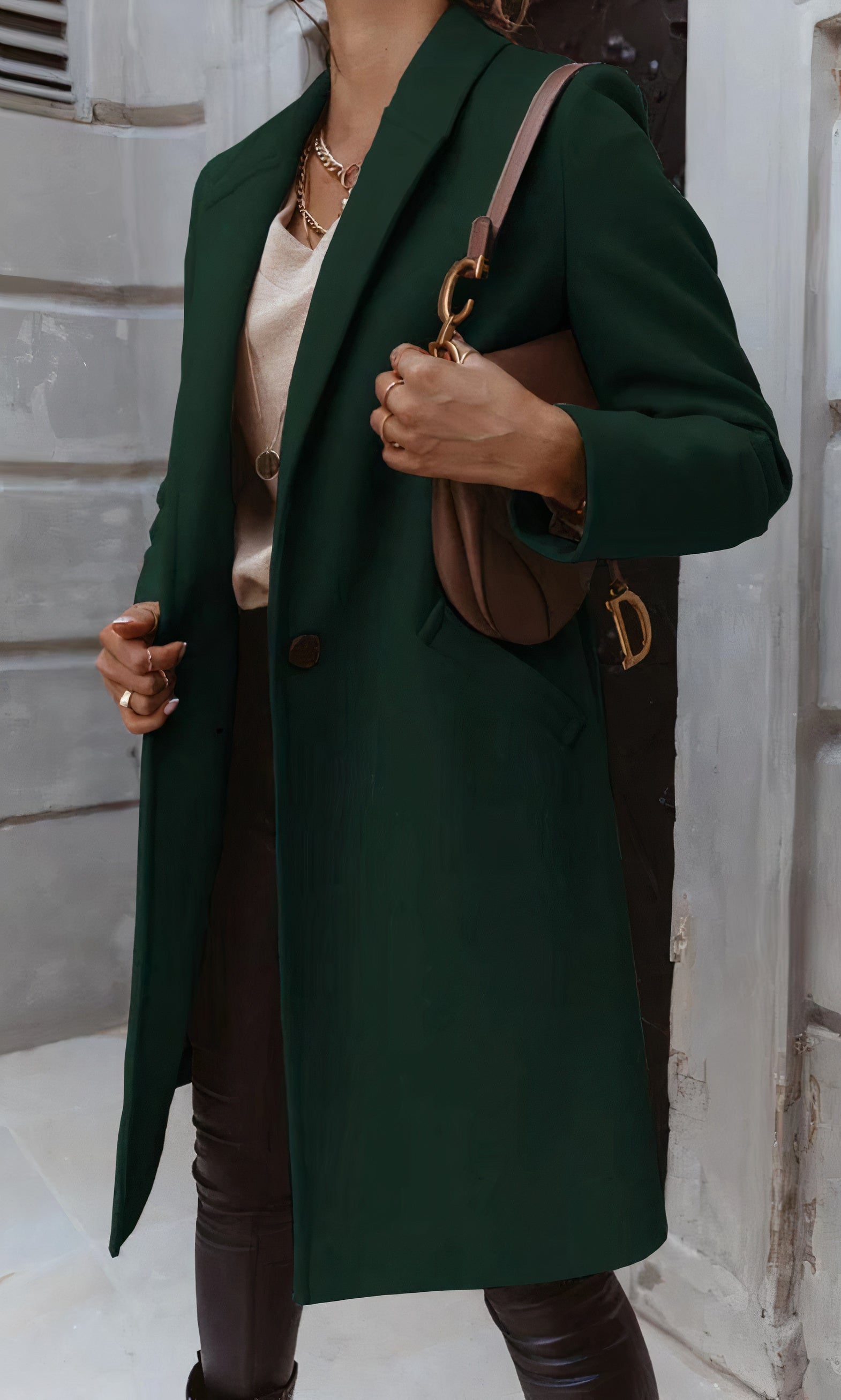 Frau in elegantem, dunkelgrünem Mantel mit brauner Umhängetasche, steht vor grauer Wand. Modetrend Herbst, stilvolle Damenbekleidung.