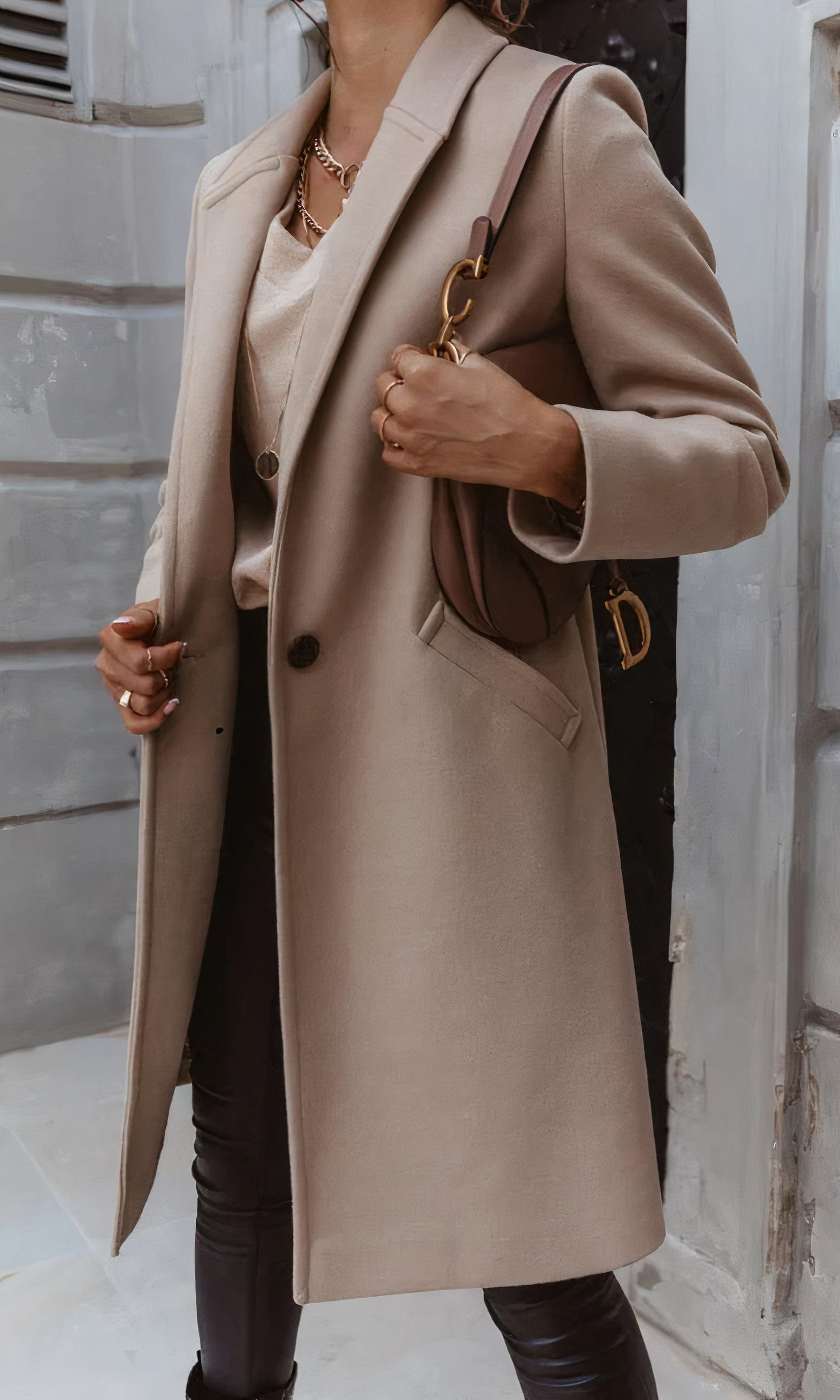 Frau in elegantem beigem Mantel, kombiniert mit brauner Tasche und schwarzer Hose, steht vor grauer Wand. Modetrend Herbst, stilvolle Damenmode.